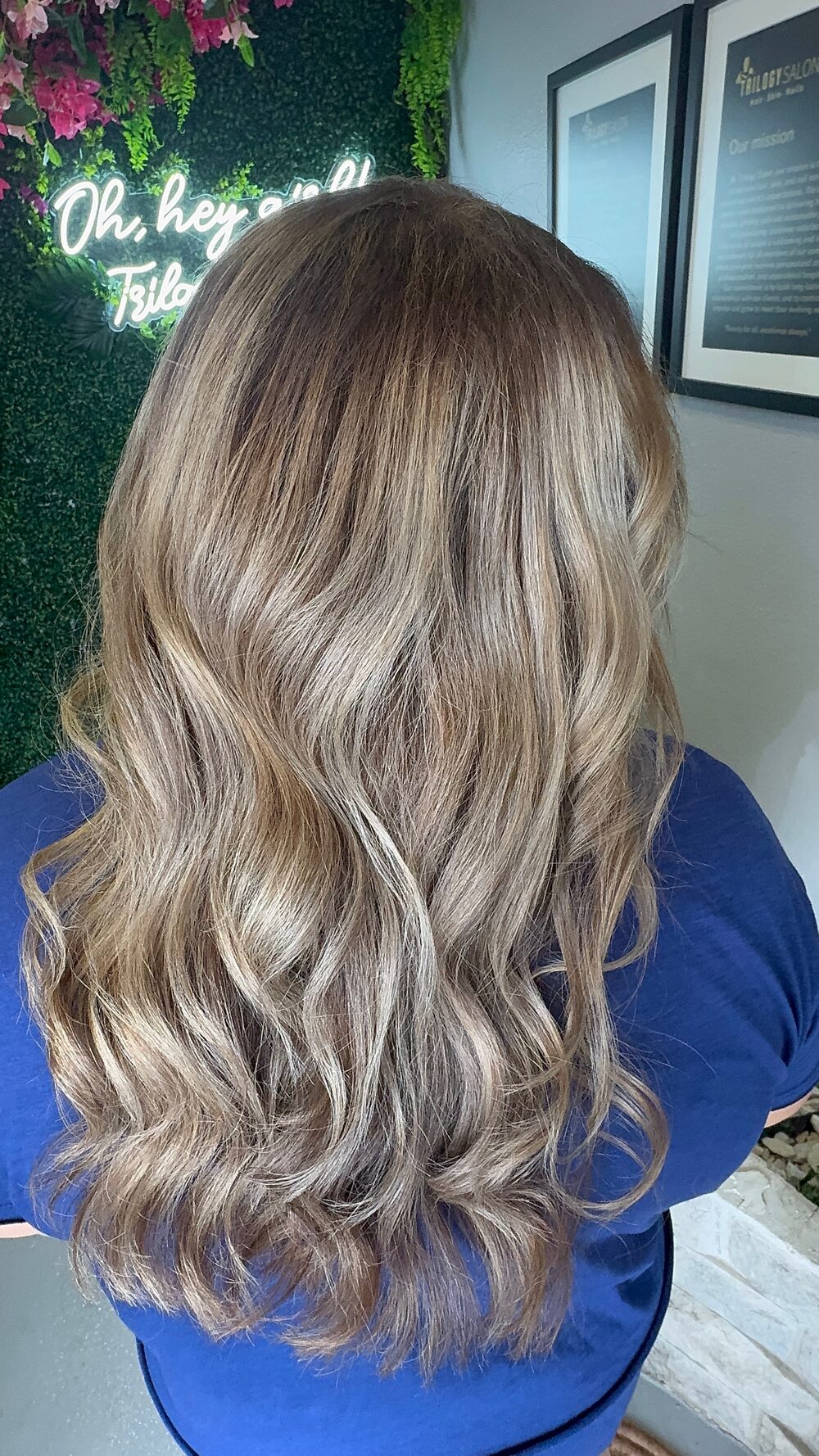 Beige blonde hair with soft waves, seen from behind in front of the salon neon sign